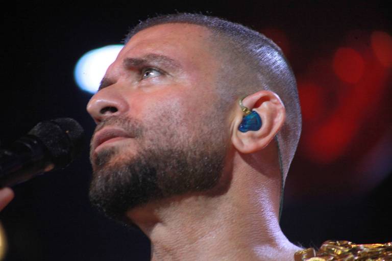 A imagem mostra um cantor em um momento de performance. Ele está de perfil, com a cabeça levemente inclinada para cima. O cantor tem cabelo curto e uma barba bem aparada. Ele usa um fone de ouvido azul e segura um microfone com a mão esquerda. O fundo é desfocado, com luzes coloridas que sugerem um ambiente de show.