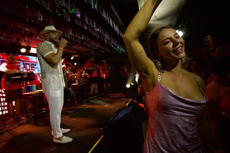 A imagem mostra um cantor em um palco, usando uma camisa brilhante e um chapéu, enquanto canta em um microfone. À sua frente, uma mulher com um vestido rosa brilhante sorri e levanta o braço, dançando. Ao fundo, há uma banda e várias pessoas assistindo ao show em um ambiente iluminado.