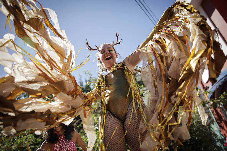 Uma mulher está em uma performance de carnaval, vestindo um traje dourado com detalhes em papel e penas. Ela tem chifres de rena e está sorrindo, com os braços abertos, criando um efeito de asas. Ao fundo, há uma outra pessoa com um vestido de bolinhas e um ambiente ao ar livre com árvores e uma casa.