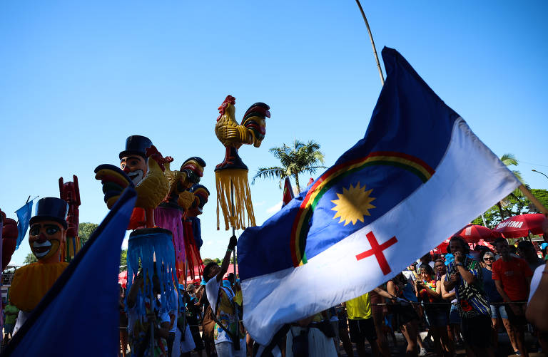 A imagem mostra uma celebração ao ar livre, com pessoas segurando bonecos grandes e coloridos, que representam figuras folclóricas. Ao centro, uma bandeira com um desenho que inclui um sol amarelo e uma cruz vermelha é visível. O céu está claro e azul, e há palmeiras ao fundo, além de uma multidão de pessoas participando do evento.