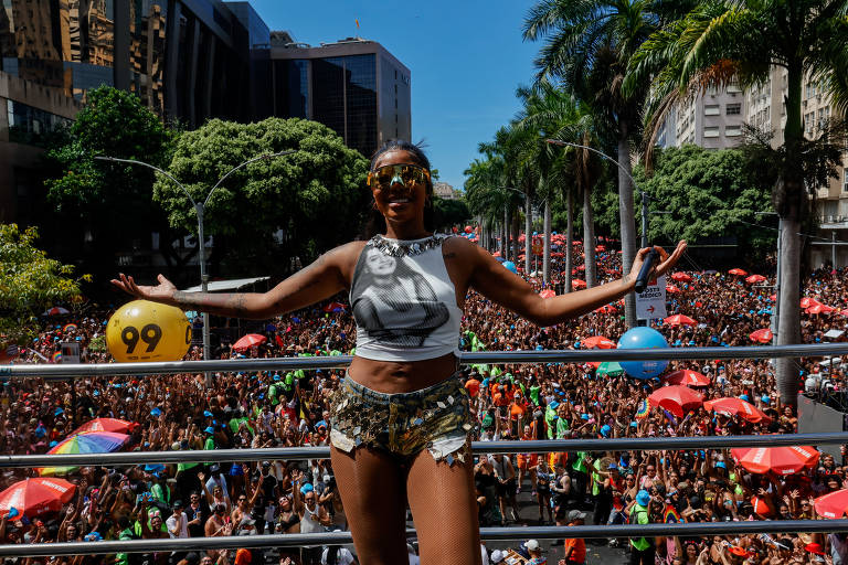 A imagem mostra uma mulher em destaque em uma plataforma elevada durante um evento de carnaval em São Paulo. Ela está vestindo uma blusa curta e shorts, com óculos de sol e sorrindo. Ao fundo, uma grande multidão de pessoas se reúne, muitas delas com guarda-sóis coloridos, sob um céu azul claro e cercada por árvores e prédios.