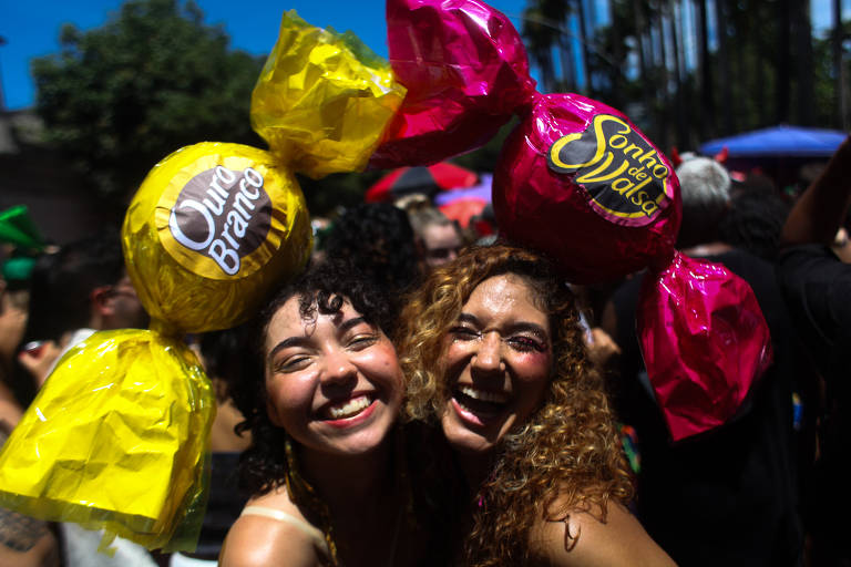 Duas mulheres sorrindo em um ambiente festivo, usando grandes chapéus em forma de doces coloridos, um amarelo e um rosa. O fundo mostra uma multidão e árvores, sugerindo um evento ao ar livre sob luz solar intensa.