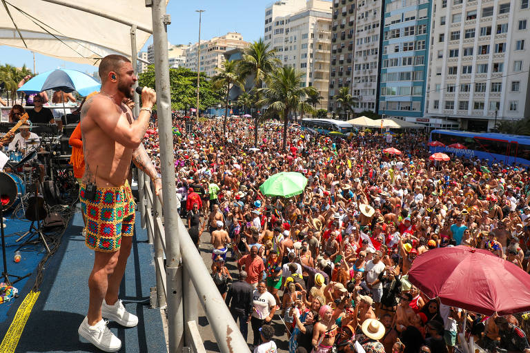 A imagem mostra uma grande multidão reunida em um evento ao ar livre, possivelmente durante o Carnaval. Um homem sem camisa, vestido com shorts coloridos, está em um palco, segurando um microfone e cantando. A multidão é composta por pessoas usando roupas de festa e chapéus, algumas segurando guarda-sóis coloridos. Ao fundo, há prédios altos e árvores, indicando um ambiente urbano ensolarado.