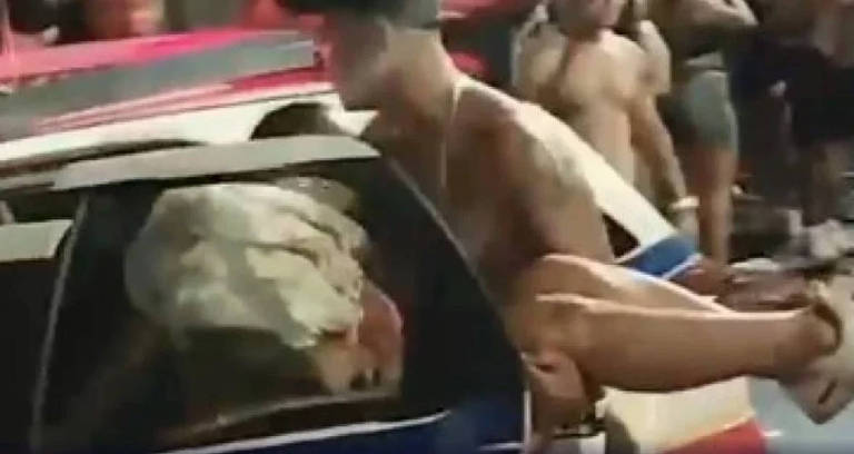 A imagem mostra uma cena em que um homem está retirando uma pessoa de dentro de um carro. O homem é musculoso e usa um boné， enquanto a pessoa que está sendo retirada parece estar inconsciente ou desmaiada. Ao fundo， há uma multidão de pessoas， algumas delas sem camisa， observando a situação. O carro é de polícia， com cores típicas de viaturas.