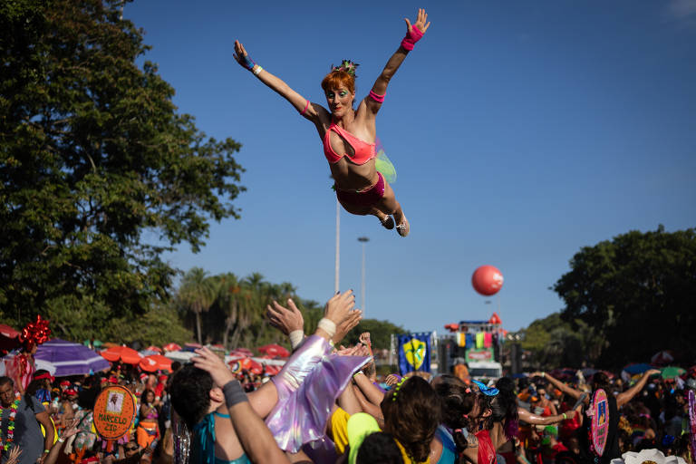 Uma mulher com cabelo laranja e fantasia colorida está sendo lançada no ar por um grupo de pessoas durante uma celebração de carnaval. O fundo mostra uma multidão animada, com árvores e barracas coloridas. O céu está claro e ensolarado.