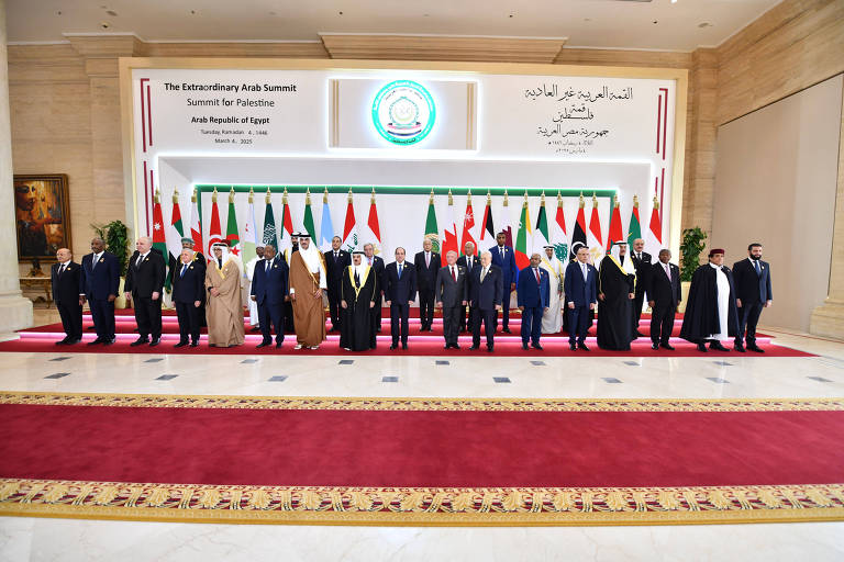 A imagem mostra um grupo de líderes árabes posando para uma foto em um evento formal. Eles estão alinhados em uma sala decorada com bandeiras de vários países árabes ao fundo. O ambiente é elegante, com um tapete vermelho e uma decoração neutra nas paredes. Há um painel com texto em árabe e inglês que indica que se trata da Cúpula Árabe Extraordinária para a Palestina, realizada na República Árabe do Egito.