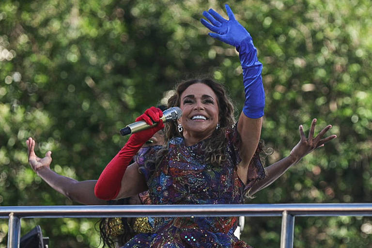 Uma artista está se apresentando em um palco ao ar livre, usando um vestido colorido e luvas de duas cores diferentes: uma vermelha e outra azul. Ela está segurando um microfone e sorrindo, enquanto gesticula com as mãos. Ao fundo, há pessoas assistindo à apresentação e uma vegetação densa.