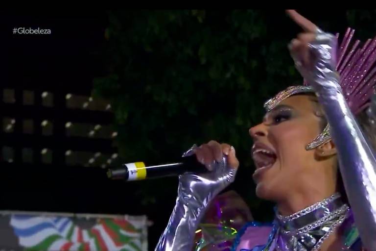 Uma artista está se apresentando em um evento ao ar livre. Ela usa um traje brilhante e colorido, com detalhes prateados e uma tiara com penas. A artista segura um microfone e parece estar cantando com entusiasmo, enquanto gesticula com a mão. Ao fundo, há uma decoração festiva.