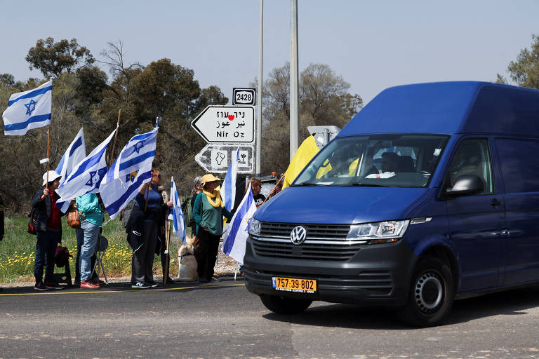 Um grupo de pessoas está em pé ao lado de uma estrada, segurando bandeiras de Israel. Elas estão vestidas com roupas coloridas e algumas usam chapéus. Um veículo utilitário azul está passando pela estrada. Ao fundo, há placas de sinalização em hebraico e árabe, indicando direções. A paisagem é composta por árvores e um campo com flores.