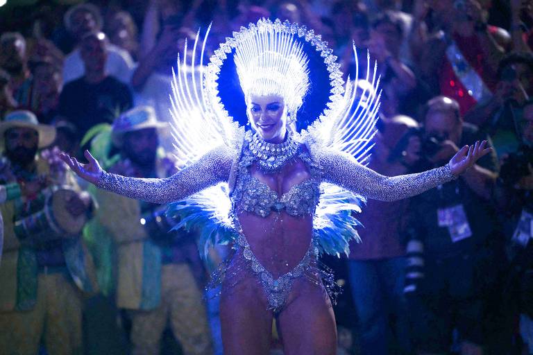 Uma mulher vestida com um traje brilhante e elaborado, adornado com penas e luzes azuis, está no centro de um desfile de carnaval. Ela tem os braços abertos, cercada por uma multidão de pessoas que a observam. Ao fundo, há músicos e foliões, todos em trajes coloridos.