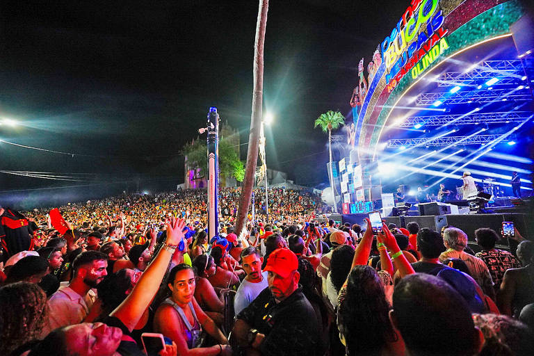 A imagem mostra uma grande multidão em um festival noturno， com pessoas levantando as mãos e se divertindo. No fundo， há um palco iluminado com luzes coloridas e um letreiro que diz 039;Carnaval039;. A atmosfera é vibrante， com muitas pessoas usando roupas festivas e algumas segurando celulares para registrar o momento.