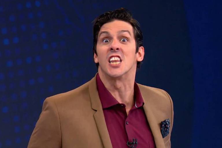 Um homem com cabelo escuro e liso, vestindo um paletó bege e uma camisa bordô, está fazendo uma expressão facial intensa, com a boca aberta e mostrando os dentes. O fundo é azul com padrões de pontos.