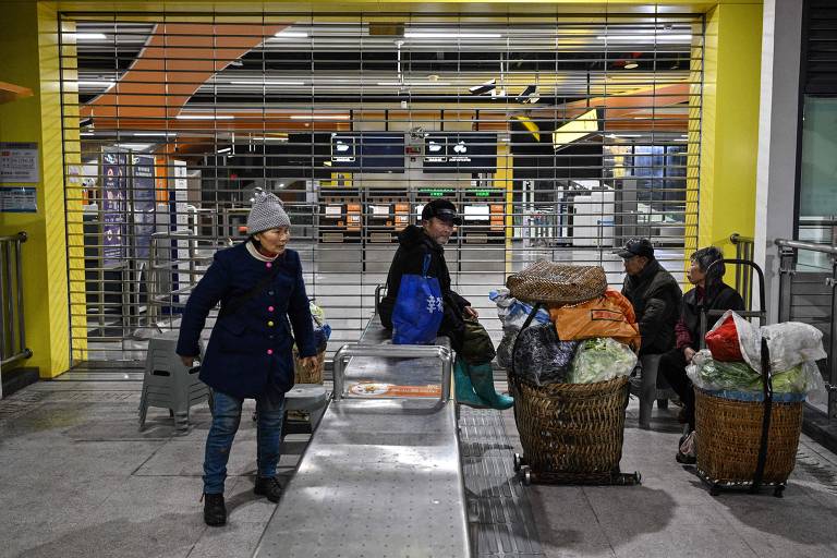 A imagem mostra uma área de uma estação de transporte com um portão de segurança fechado ao fundo. À esquerda, uma mulher com um gorro e um casaco azul está em pé, enquanto um homem sentado à direita usa um chapéu e está cercado por cestos de vime cheios de produtos. Outros dois homens estão sentados em bancos, um deles com uma bolsa ao lado. O ambiente é iluminado, mas parece ser de noite.