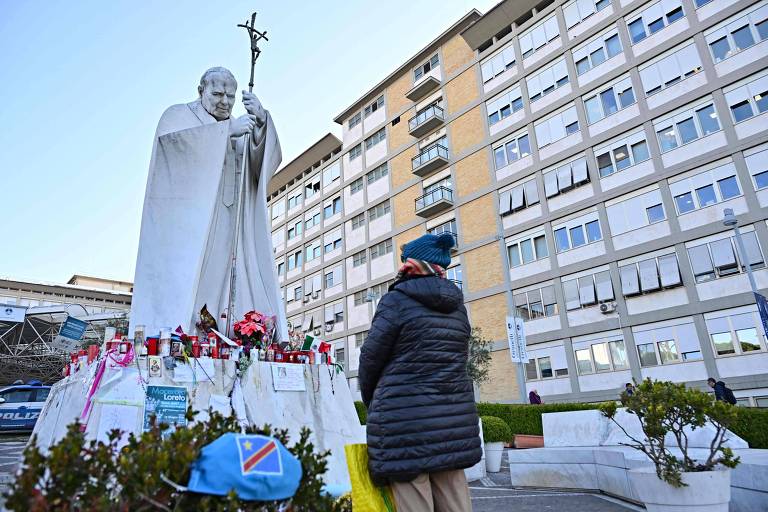 A imagem mostra uma estátua de Papa João Paulo II, vestindo uma túnica e segurando uma cruz. A estátua está cercada por flores, velas e outros tributos. À frente da estátua, uma pessoa está de costas, usando um casaco escuro e um gorro azul. Ao fundo, há um edifício de apartamentos com janelas e varandas.