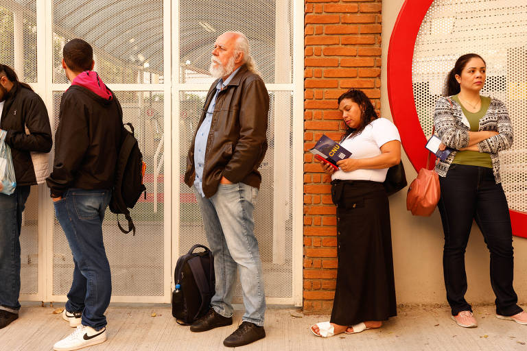 A imagem mostra um grupo de cinco pessoas em uma fila, em um ambiente urbano. À esquerda, um homem jovem com um moletom preto e capuz está olhando para o celular. Ao lado dele, um homem mais velho com cabelo e barba brancos, vestindo uma jaqueta de couro, está em pé, observando a fila. Uma mulher de cabelo cacheado, vestindo uma camiseta branca e uma saia longa preta, está segurando um documento. À direita, uma mulher com cabelo preso, usando uma blusa verde e um casaco de malha, está de braços cruzados. A fila está em frente a uma parede com uma grade metálica e tijolos expostos.