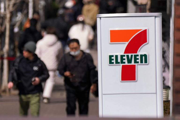 A imagem mostra um sinal da loja 7-Eleven em primeiro plano， com o número 7 em laranja e verde， e a palavra 039;ELEVEN039; em verde. Ao fundo， há pessoas caminhando em uma rua movimentada， algumas usando máscaras e vestindo roupas de inverno.