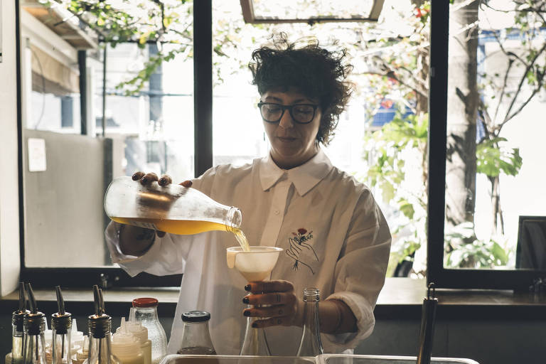 Uma mulher com cabelo cacheado e óculos está em um bar, despejando uma bebida de uma jarra em um copo. A pessoa usa uma camisa branca e está cercada por garrafas e recipientes de ingredientes. Ao fundo, há uma grande janela com vista para árvores e luz natural entrando.