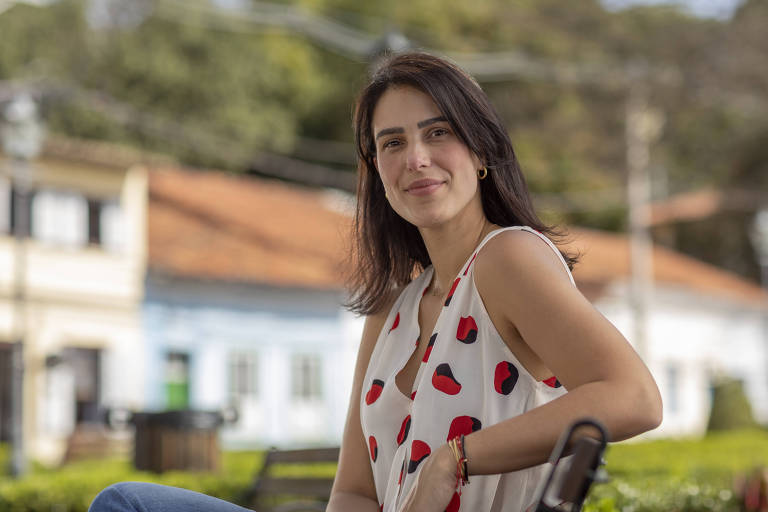 Uma mulher está sentada em um parque, sorrindo para a câmera. Ela usa uma blusa branca com estampas de morangos e tem cabelo solto. Ao fundo, há casas com telhados de barro e árvores, criando um ambiente natural e tranquilo.