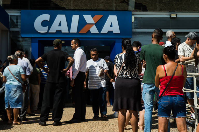 Uma fila de pessoas se forma em frente a uma agência da Caixa Econômica Federal. O letreiro da agência é visível, destacando o nome 'CAIXA' em letras grandes. As pessoas na fila estão vestidas de maneira casual, algumas usando camisetas e outras com roupas de verão. O ambiente parece ser ensolarado e há uma mistura de homens e mulheres na fila.