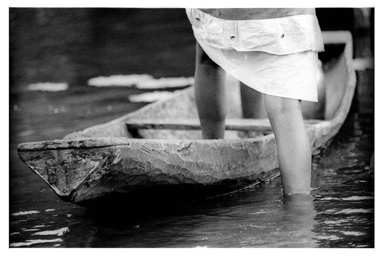 A imagem em preto e branco mostra uma pessoa de pernas nuas em pé dentro de uma canoa de madeira, com água ao redor. A parte inferior da canoa é visível, e a água parece calma. A pessoa está vestindo uma saia branca que flutua levemente na água.