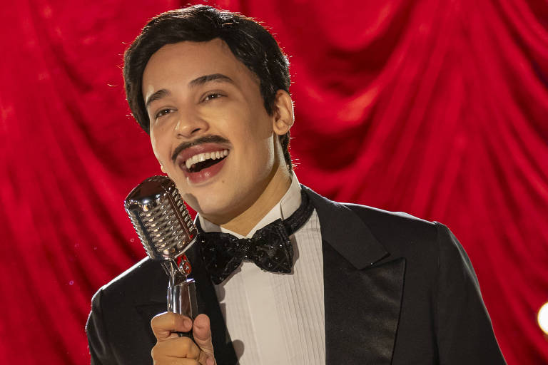 Um homem em um terno preto com gravata borboleta está em um palco, segurando um microfone vintage. Ele tem um bigode e está sorrindo, parecendo se divertir enquanto canta. O fundo é composto por cortinas vermelhas e há algumas luzes de palco acesas.