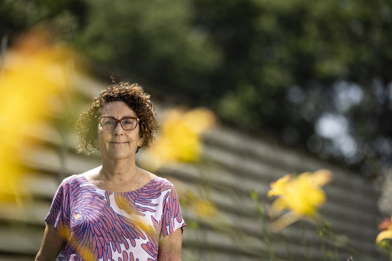 A imagem mostra uma mulher de pé em um ambiente ao ar livre, com flores amarelas em primeiro plano. Ela tem cabelo cacheado e usa uma blusa com estampas em tons de roxo e branco. Ao fundo, há uma cerca de madeira e árvores desfocadas.