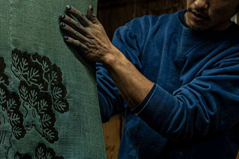 Um homem está manipulando um grande pedaço de tecido verde, que possui um padrão de árvore escura. Ele usa uma camiseta azul e tem as unhas pintadas de preto. O ambiente parece ser um ateliê, com iluminação suave e um fundo de madeira.