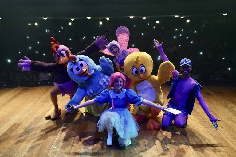 Um grupo de seis personagens fantasiados em um palco. Os personagens incluem aves coloridas e uma figura central com cabelo rosa e vestido azul. O fundo é escuro, com luzes brilhantes de celulares na plateia, criando um ambiente festivo.