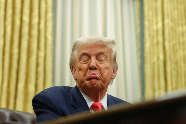 A imagem mostra um homem com cabelo loiro e uma expressão facial de descontentamento, sentado em um ambiente formal. Ele está usando um terno escuro e uma gravata vermelha. O fundo apresenta cortinas douradas e uma mesa escura à frente.