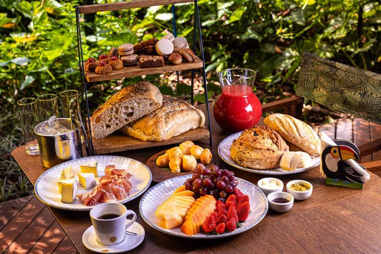 A mesa apresenta um café da manhã variado com pães, frutas e bebidas. No centro, há uma bandeja de dois tipos de pães, acompanhada de uma jarra de suco vermelho. Ao redor, estão pratos com frutas como melão, morango e uvas, além de fatias de queijo e presunto. Também há uma xícara de café e um pequeno recipiente com manteiga. O fundo é composto por uma vegetação verde.