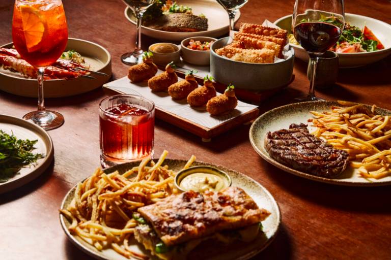 A imagem mostra uma mesa de jantar com diversos pratos de comida. Há um prato com batatas fritas e um filé grelhado, um prato com um peixe grelhado, e outro com croquetes. Também estão presentes copos com bebidas, incluindo um coquetel colorido e um copo de bebida escura. Ao fundo, há mais pratos e saladas.