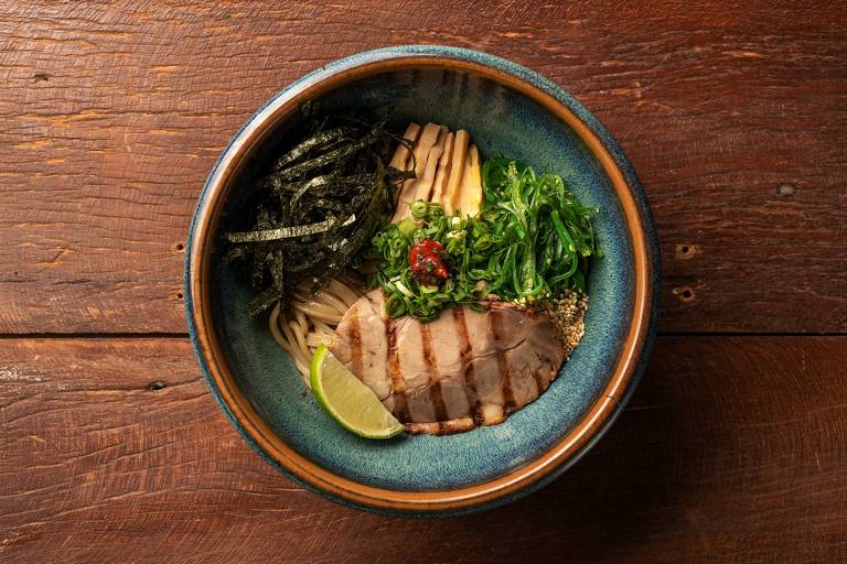 Uma tigela de ramen com fatias de carne grelhada, algas marinhas, cebolinhas, um limão cortado e um toque de pimenta vermelha. O fundo é de madeira, e a tigela é de cerâmica azul com borda marrom.