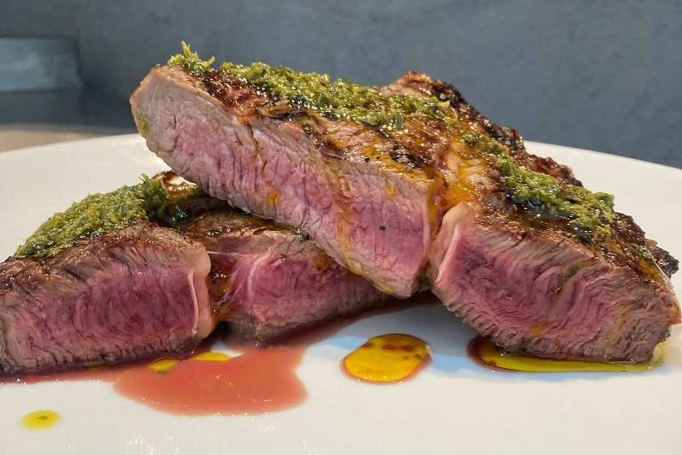 A imagem mostra três pedaços de carne grelhada dispostos em um prato branco. A carne apresenta uma crosta verde em cima e é cortada ao meio, revelando um interior rosado. Ao redor da carne, há manchas de molho vermelho e amarelo.