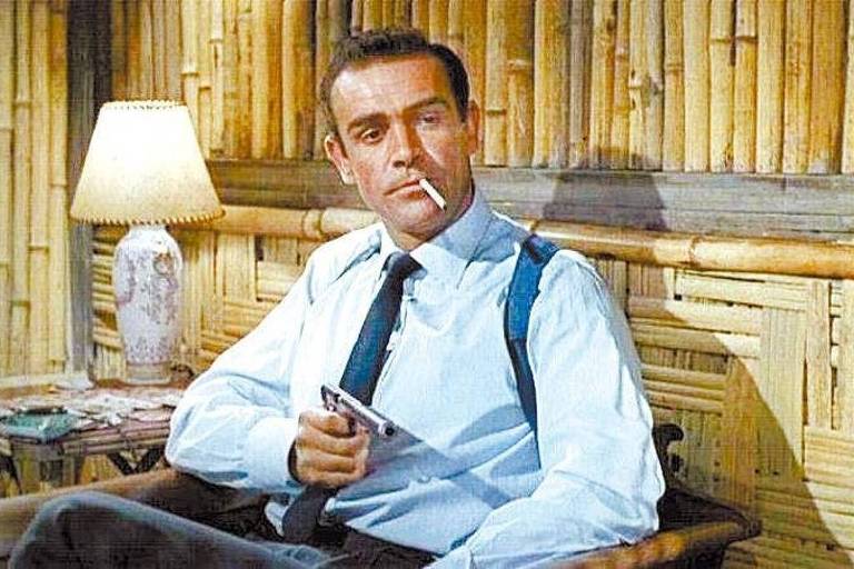 Um homem, o ator Sean Connery, está sentado em uma cadeira em um ambiente com decoração de bambu. Ele está usando uma camisa social clara e gravata, com suspensórios visíveis. O homem segura um cigarro na boca e uma arma na mão. Ao fundo, há uma lâmpada acesa em uma mesa ao lado, e a mesa tem alguns objetos em cima, entre eles um abajur.
