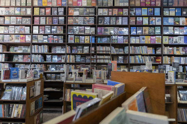 A imagem mostra o interior de uma livraria, com estantes repletas de livros organizados em várias prateleiras. No primeiro plano, há mesas ou estantes baixas com livros visíveis. As prateleiras ao fundo estão cheias de capas de livros de diferentes cores e tamanhos, criando um ambiente acolhedor e convidativo para os leitores.