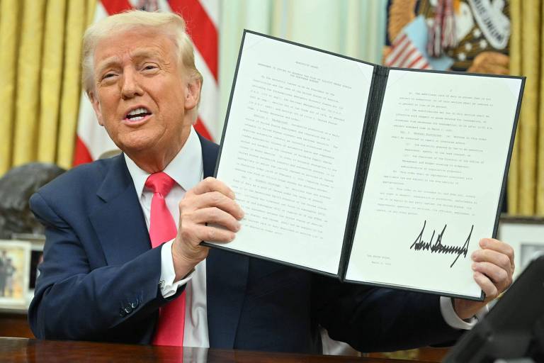 Um homem está sentado em uma mesa, segurando um documento com uma assinatura visível. Ele está sorrindo e vestindo um terno escuro com uma gravata rosa. Ao fundo, há bandeiras e outros itens decorativos.