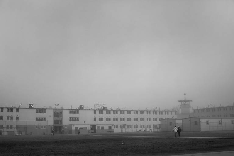 A imagem em preto e branco mostra uma prisão em um dia nublado. O edifício da prisão é de vários andares, com janelas visíveis e cercas de segurança no topo. No primeiro plano, duas pessoas caminham em direção ao edifício, enquanto o céu está coberto por uma névoa densa