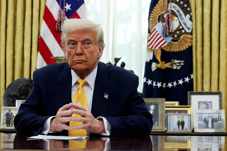 Um homem sentado em uma mesa no Escritório Oval, com bandeiras dos Estados Unidos ao fundo. Ele tem cabelo loiro e usa um terno escuro com uma gravata amarela. A mesa está decorada com várias fotos em molduras e um objeto decorativo à esquerda.