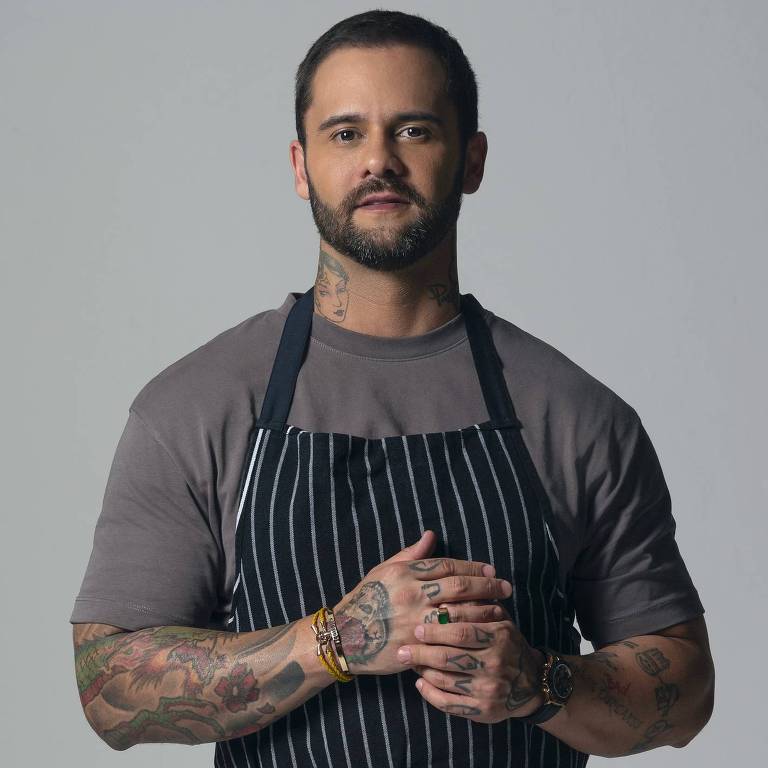 Um homem com cabelo curto e barba, vestindo uma camiseta cinza e um avental listrado preto e branco. Ele está de pé, com as mãos unidas na frente do corpo, em um fundo cinza claro.