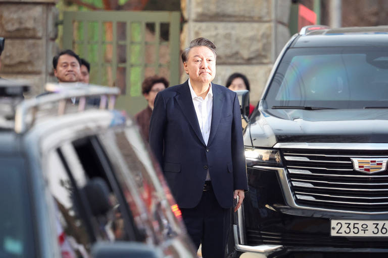 Um homem em traje formal, com um paletó escuro e camisa clara, está em pé ao lado de um veículo grande, possivelmente um SUV. Ao fundo, há várias pessoas observando, algumas sorrindo. O ambiente parece ser uma área urbana, com uma entrada visível ao fundo.