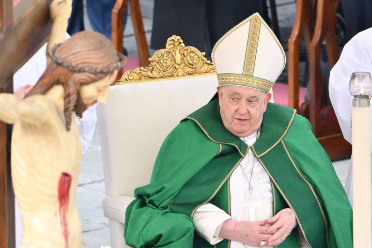 A imagem mostra um líder religioso sentado em uma cadeira, vestindo uma túnica verde e um chapéu papal. Ele está em frente a uma mesa com um altar, onde há uma cruz com a figura de Jesus crucificado. Ao fundo, há outros clérigos e elementos decorativos como velas. A cena parece ser parte de uma cerimônia religiosa ao ar livre.