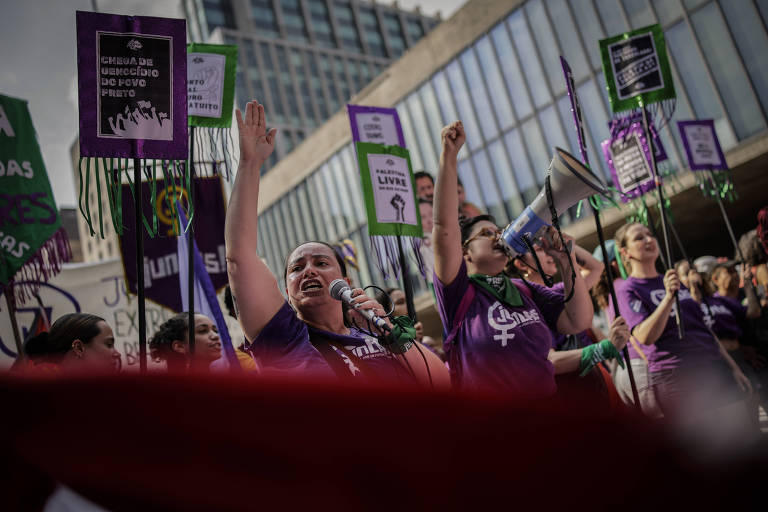 A imagem mostra um grupo de mulheres participando de uma manifestação. Elas estão vestidas com camisetas roxas e seguram cartazes com mensagens de empoderamento e direitos femininos. Uma mulher à frente está usando um microfone, enquanto outra segura um megafone. O ambiente é urbano, com prédios ao fundo.