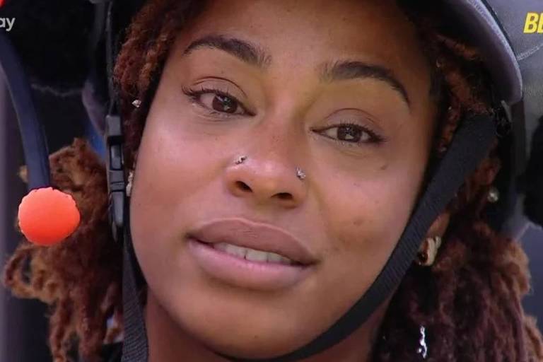 A imagem mostra um close-up de uma mulher com cabelo cacheado e dreadlocks. Ela está usando um capacete e fones de ouvido, com um olhar sério. A iluminação é suave, destacando suas características faciais, como piercings no nariz e nas orelhas.