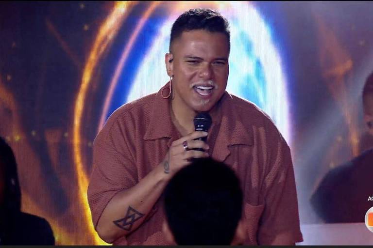 Um artista está se apresentando ao vivo em um palco iluminado. Ele usa uma camisa de cor clara e está segurando um microfone, sorrindo enquanto canta. Ao fundo, há outros músicos e uma iluminação vibrante que cria um efeito visual dinâmico.