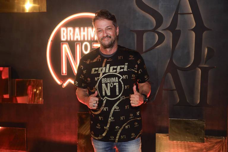 Um homem está posando para a foto em um evento. Ele usa uma camiseta preta com estampas douradas que incluem o logo da Brahma N°1. O fundo apresenta uma iluminação suave e elementos decorativos em dourado e preto, com a palavra 'SÃO PAULI' visível.