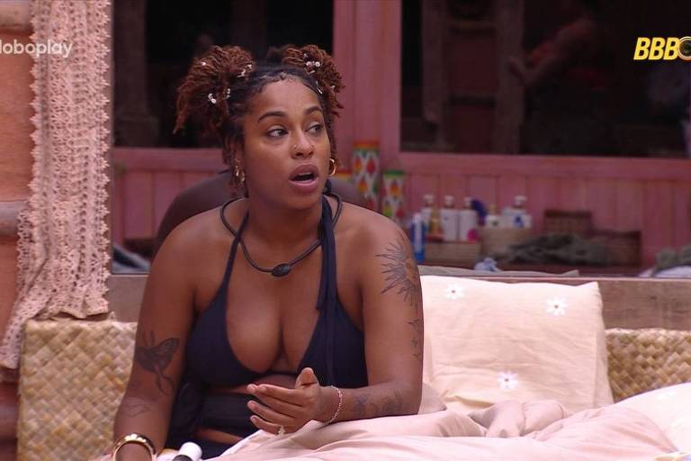 Uma mulher com cabelo crespo e preso em dois coques, usando um top preto, está sentada na cama. Ela parece estar em uma conversa, com uma expressão de surpresa ou preocupação. O ambiente ao redor é decorado com paredes de tom rosa e objetos pessoais visíveis ao fundo.