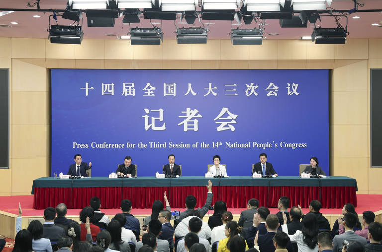 A imagem mostra uma conferência de imprensa com um painel de seis pessoas sentadas em uma mesa. Ao fundo, há um grande banner azul com texto em chinês e a frase em inglês 'Press Conference for the Third Session of the 14th National People's Congress'. A audiência, composta por várias pessoas, está visível na parte inferior da imagem, algumas levantando as mãos.