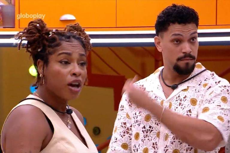 A imagem mostra duas pessoas em um ambiente interno colorido. A mulher à esquerda tem cabelo crespo preso em coquinhos e usa uma blusa sem mangas de cor clara. O homem à direita tem cabelo curto e barba, vestindo uma camisa branca com detalhes em crochê. Ambos parecem estar em uma conversa, com expressões faciais que indicam atenção.
