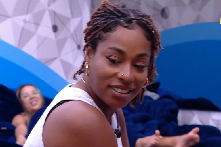 A imagem mostra uma mulher com cabelo cacheado e dreadlocks, vestindo uma blusa branca, olhando para o lado com um sorriso. Ao fundo, outra mulher está deitada em um sofá, visível parcialmente, com os pés descalços. O ambiente é decorado em tons de azul e possui um design moderno.