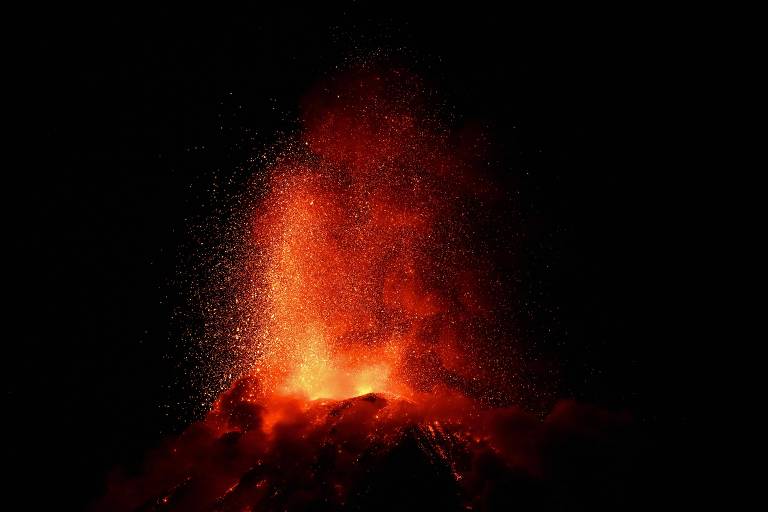 A imagem mostra uma erupção vulcânica com lava e cinzas sendo expelidas do topo de um vulcão. O fundo é escuro, destacando a luminosidade da lava e as partículas que estão sendo lançadas para o ar.